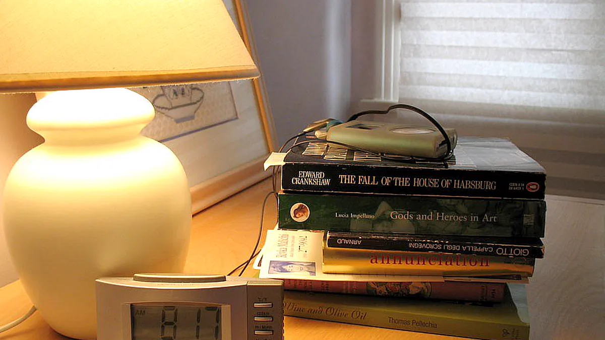 Nightstand with a glowing ceramic table lamp, a stack of hardcover books and a digital alarm clock by a window blind