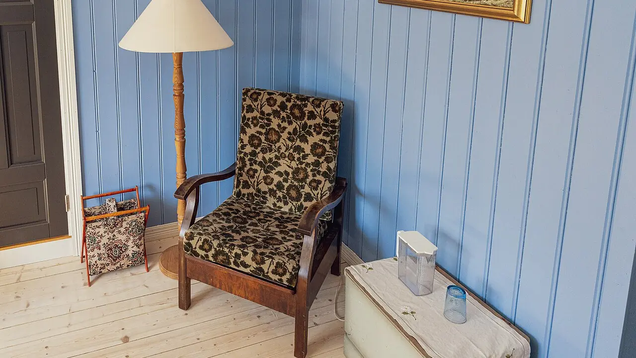 An upholstered armchair beside a floor lamp and a magazine rack in a corner with blue wooden walls