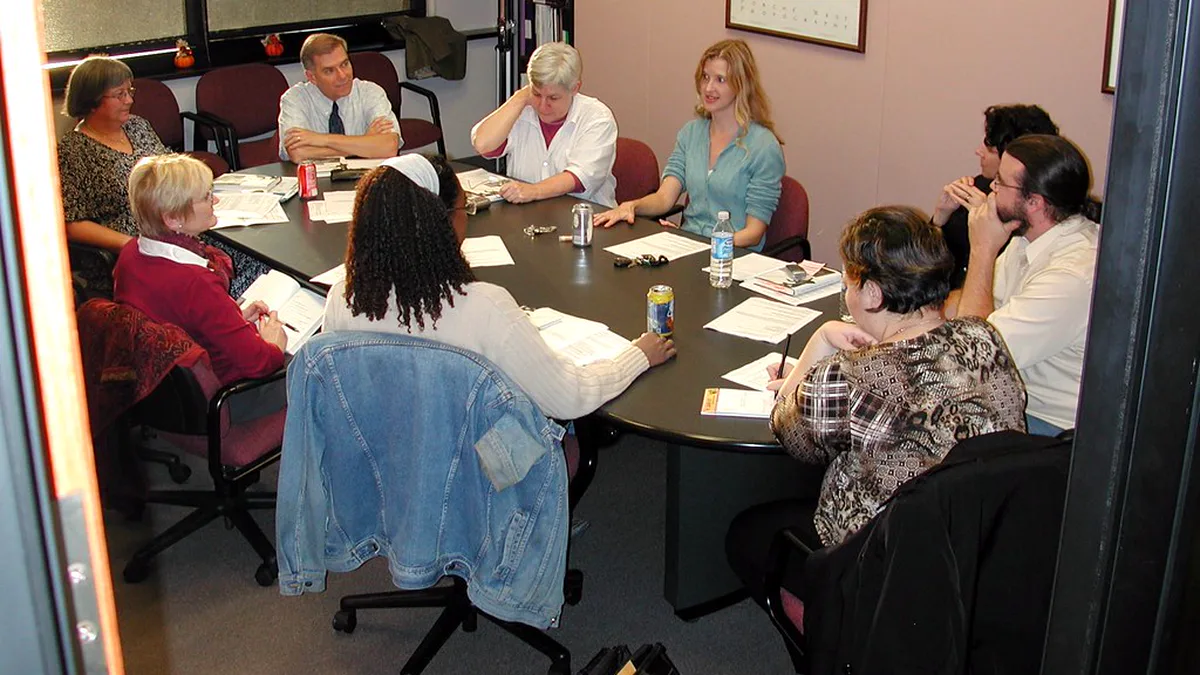 Book Discussion Group Around One Table With Printed Notes Ten people seated around a dark round meeting table with printed notes and drinks, mid-conversation at a book club meeting