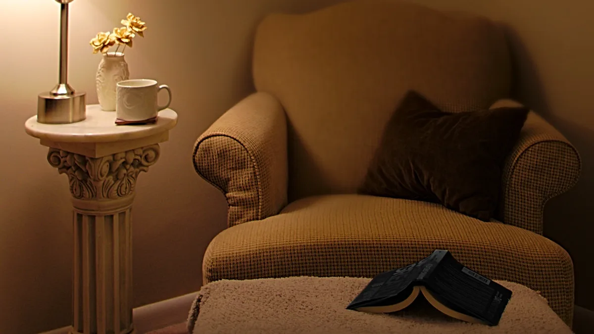 Cozy reading nook in the evening with an upholstered armchair, a lamp and mug on a pedestal side table and an open book on a footstool