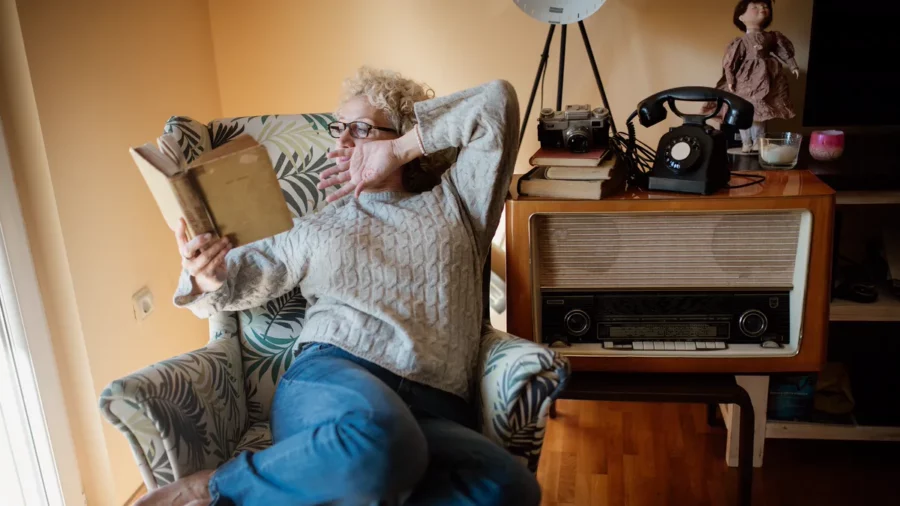 Woman in a grey sweater reading a paperback in a patterned armchair by a sunny window, a way to get back into reading at home