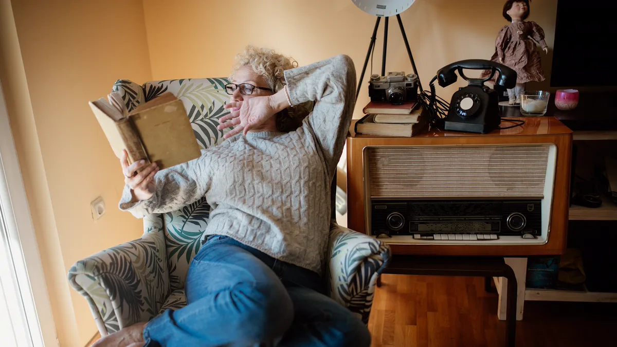 Reader Stretching in an Armchair by the Window With a Paperback Woman in a grey sweater reading a paperback in a patterned armchair by a sunny window, a way to get back into reading at home