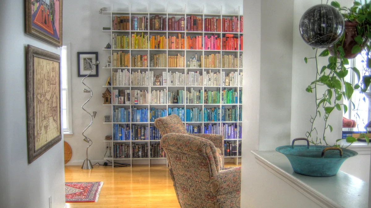 Color-Sorted Home Library Bookcase Tall white bookcase sorted by spine color in a sunlit living room, showing how to organize a bookshelf as a rainbow
