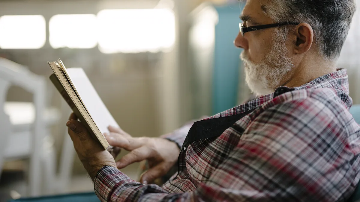 Grey-haired man in glasses and a plaid shirt reading a paperback in an armchair, steady practice being how to read faster