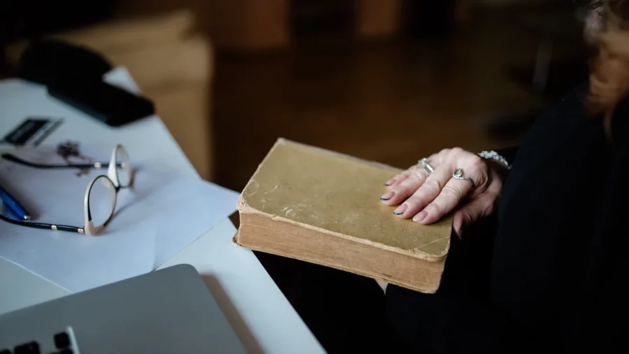 A reader rests one hand on a worn cloth-bound book beside a desk with reading glasses, papers and a laptop, pausing to recall what they read