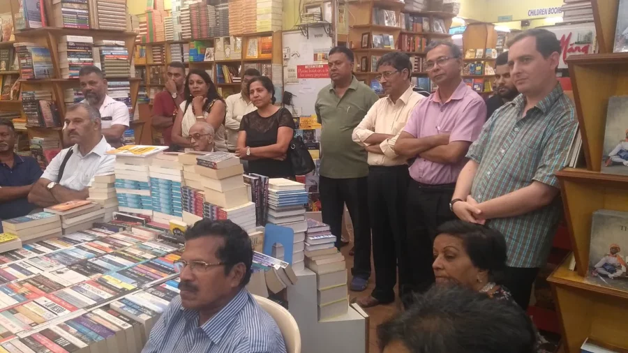 A group of adult readers sitting and standing between crowded bookshop shelves and stacked paperbacks at a book club meeting