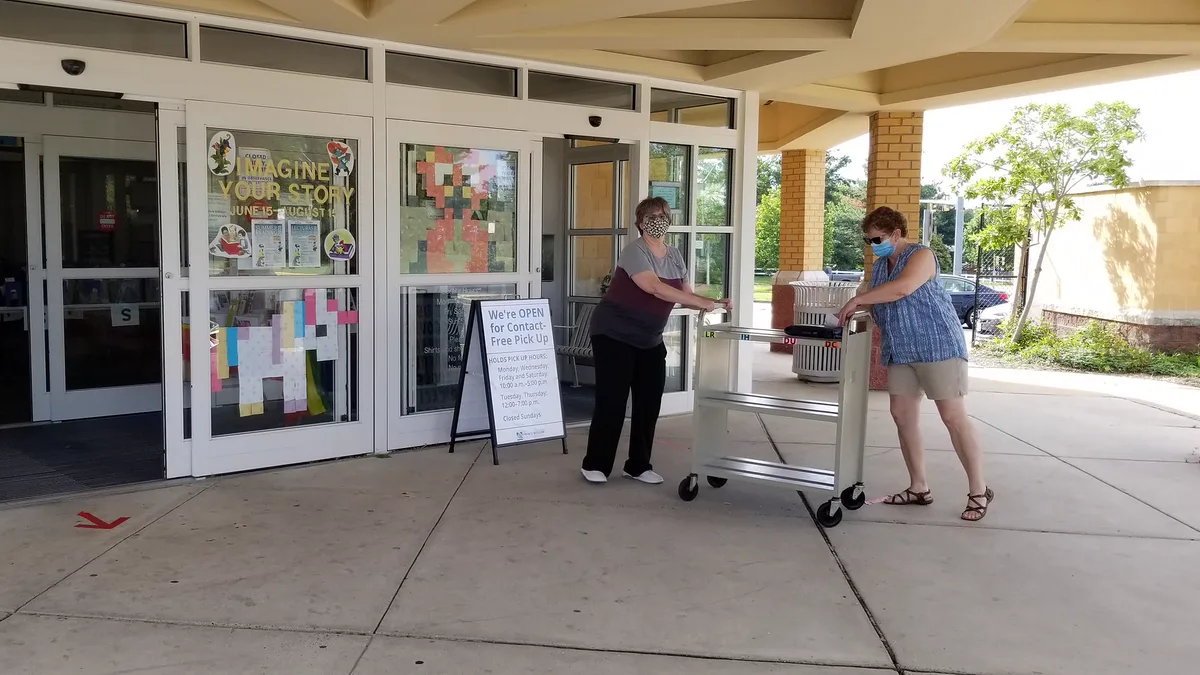 Library Staff Bringing Out Reserved Books for Pickup Two library staff members wheel a book cart out of the Bull Run Library entrance to hand reserved hold books to patrons