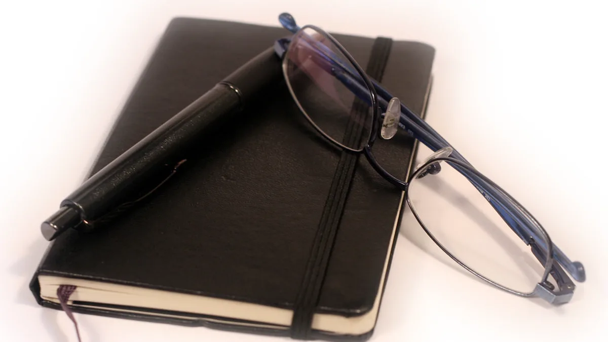 Notebook, Pen and Glasses for a One-Line Book Summary A black elastic-band notebook with a pen and a pair of reading glasses resting on top, ready for a short summary after a reading session