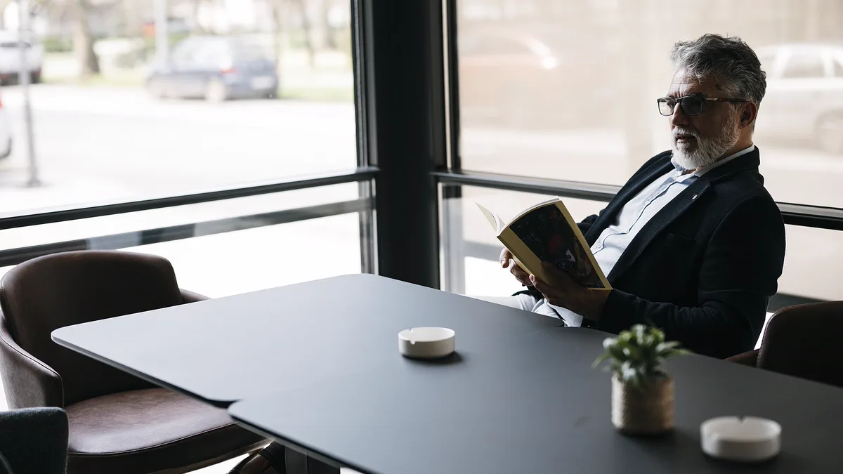 Man in a dark jacket reading a paperback at an empty cafe table beside a large window, using a spare slot to read more