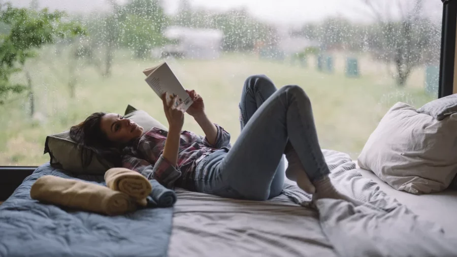 Woman lying on a bed with her knees up, reading a paperback beside a large window on a rainy evening, a calm reading before bed moment