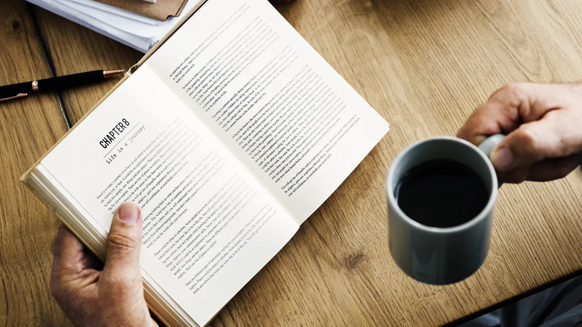 Open Paperback and a Mug of Coffee Hands holding an open paperback and a mug of black coffee at a table, a quiet seasonal reading moment