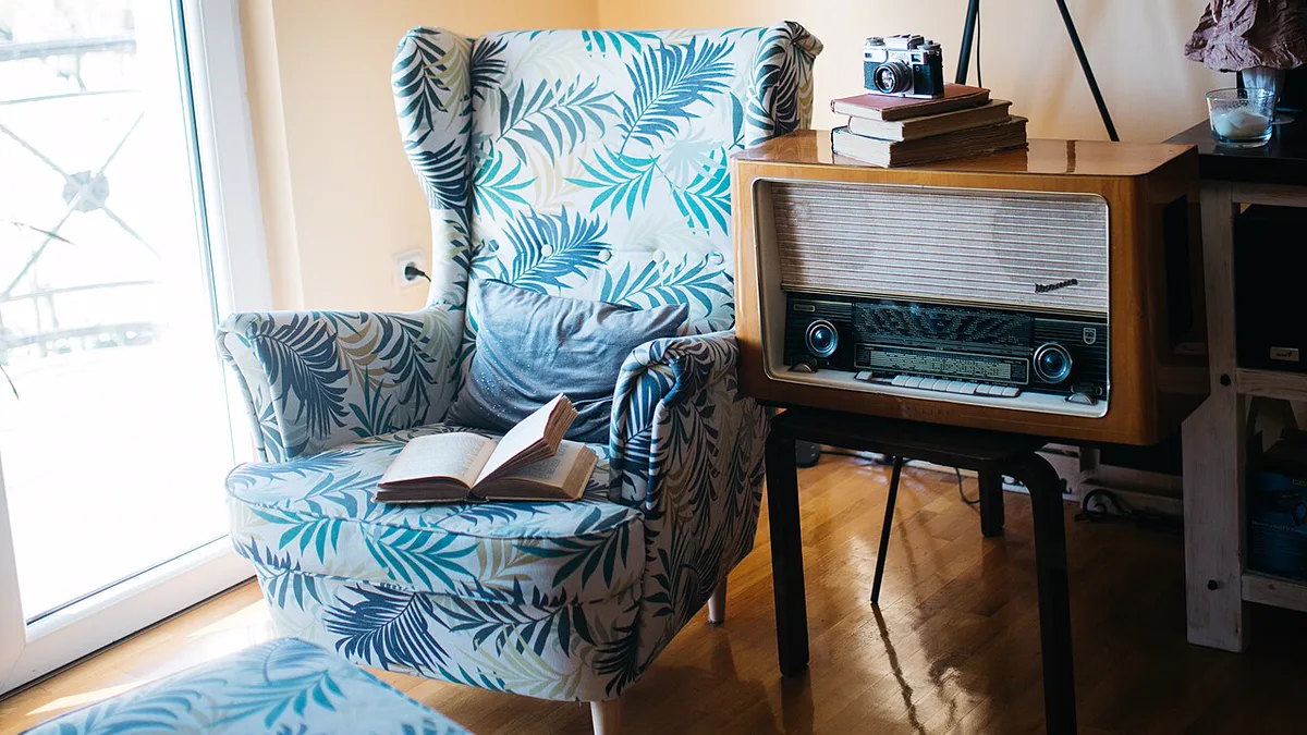 Small reading corner beside a balcony door with a leaf print wingback chair, matching ottoman, an open book and a tripod lamp