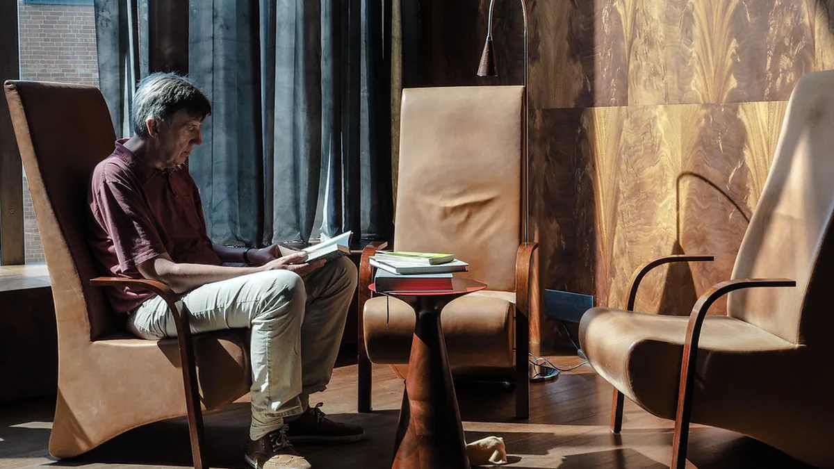 Reader in an Armchair by the Window Man reading a book in a wooden armchair near tall curtains, the kind of quiet daily session a minutes-based reading goal counts