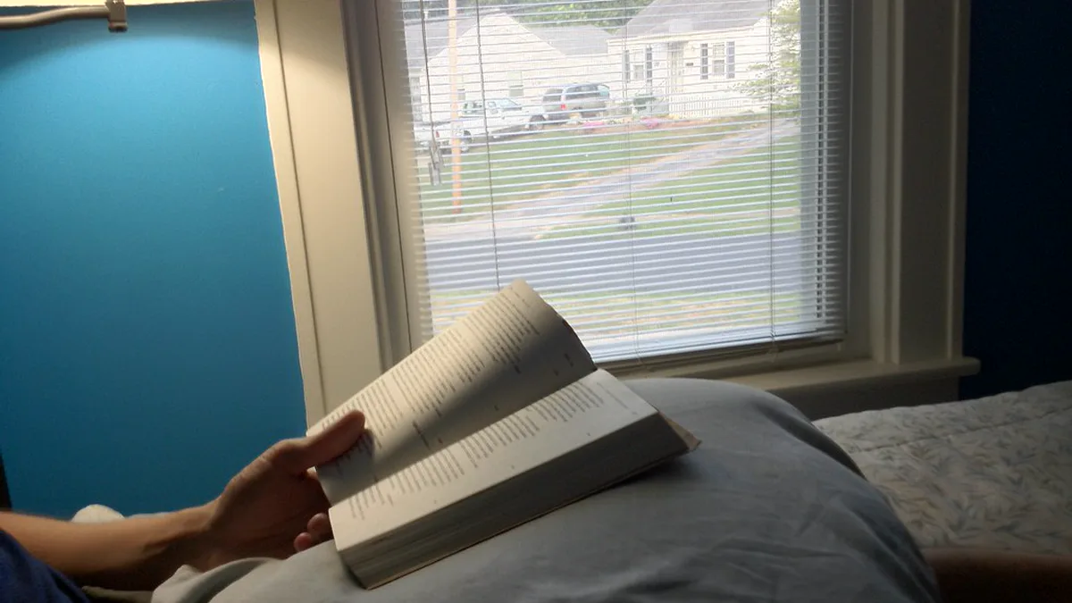 Hand holding an open paperback over a gray duvet while reading in bed, with a lamp and a window with blinds behind