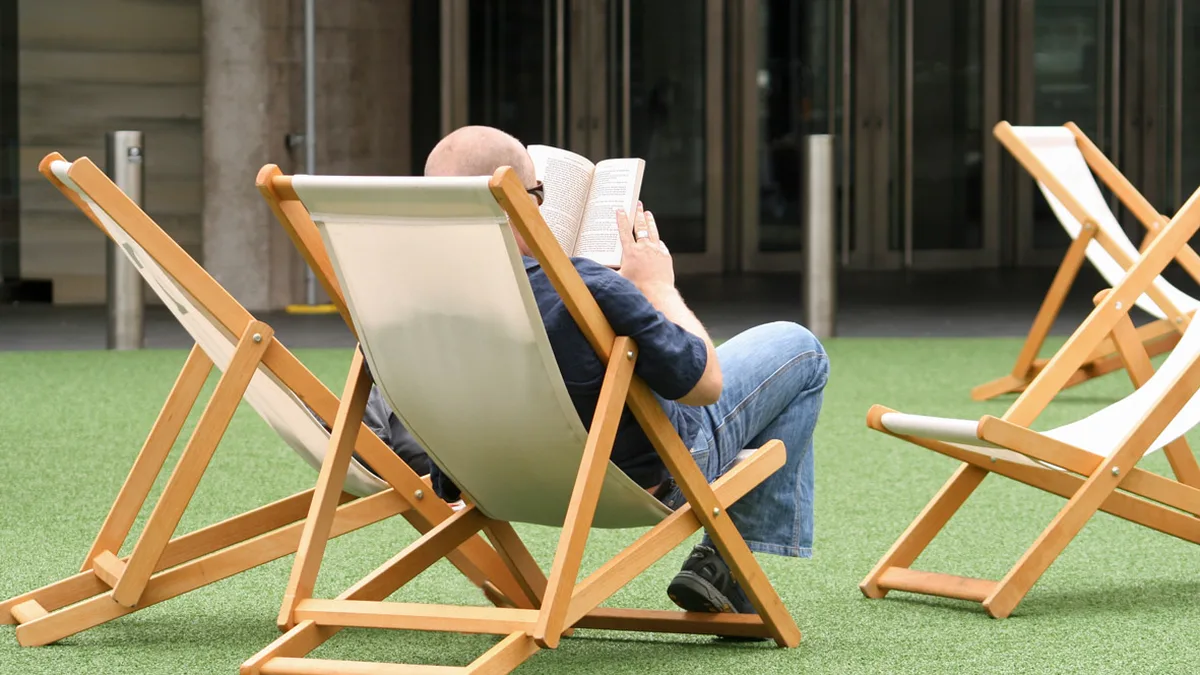 Reader in a Deck Chair Deciding Whether to Keep Going A man seen from behind reads a paperback in a wooden deck chair on artificial grass, deciding whether to finish a book he isn't enjoying